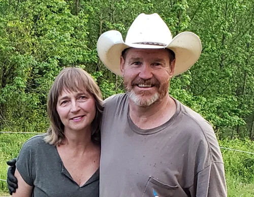 A smiling couple poses outdoors, surrounded by lush green trees. The woman has shoulder-length hair and wears a gray top, while the man, wearing a white cowboy hat and a gray T-shirt, stands beside her with a friendly expression.