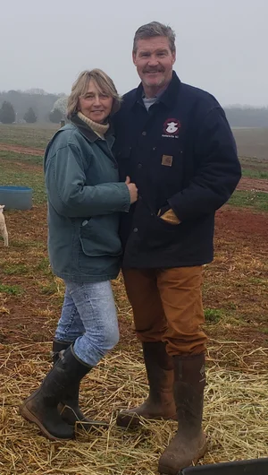 Couple standing together in a foggy farm setting, wearing casual outdoor clothing and rubber boots, with farmland in the background.